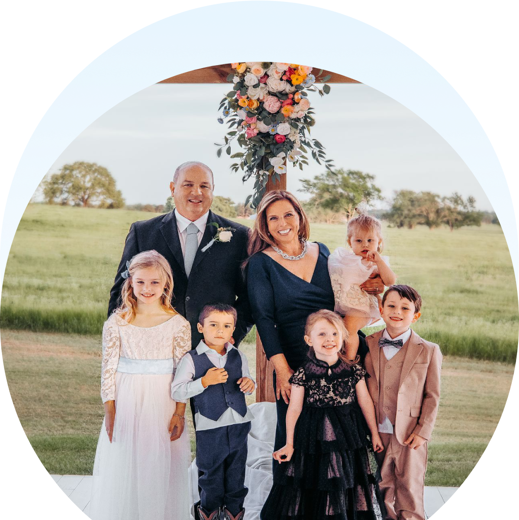 A couple and five children pose under a floral arch outdoors. The adults are dressed in formal wear, and the children are wearing a mix of dresses and suits. A grassy field is visible in the background.